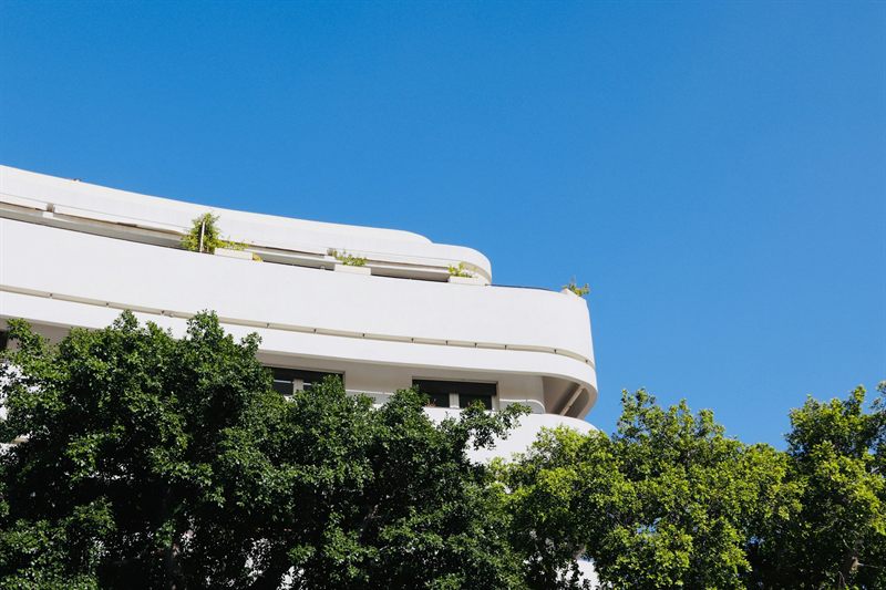 A white Bauhaus building with rounded balconies above green trees in Tel Aviv